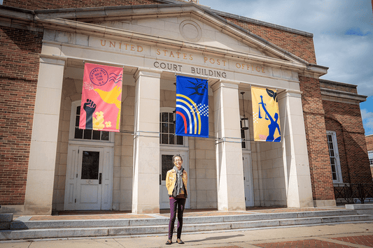 Artist Victoria Primicias standing in front of her "New Voices" banner artwork at the Peace & Justice Plaza, Chapel Hill.